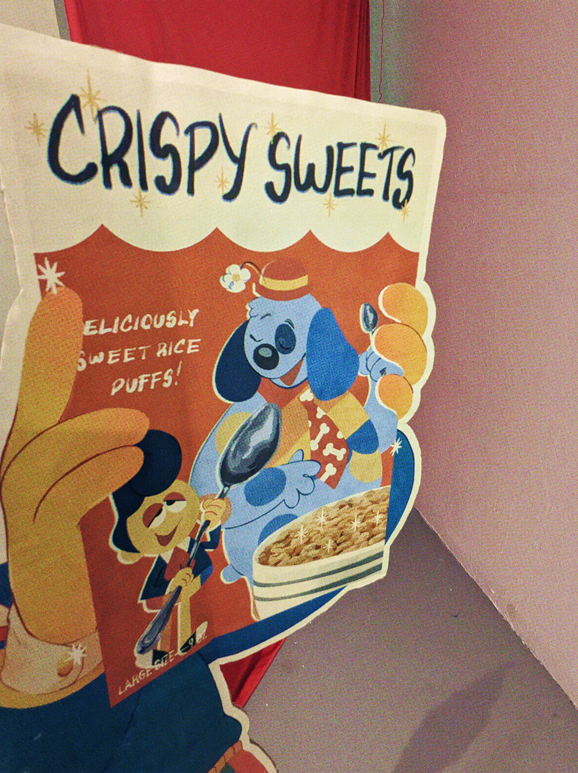 A phorograph of a cardboard standee illustrated to have Wally Darling holding a box of Crispy Sweets, Wally and Barnaby are on the box, and it has the slogan 'Deliciously Sweet Rice Puffs' on it